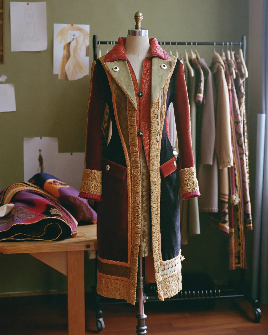 Un costume di scena in velluto e broccato montato sul manichino, davanti a uno stand di capi e ai bozzetti appesi